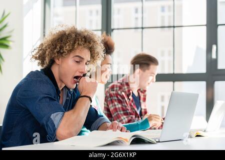 Side view portrait of a funny student yawning in front of a book, while sitting down at desk in the classroom at a modern college or university Stock Photo