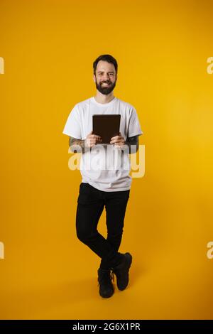 Happy mid aged brunette bearded man holding digital tablet standing over yellow wall Stock Photo