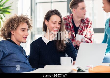 Two young students searching for an answer to their question both online and in a textbook while sitting at desk in the classroom during break Stock Photo
