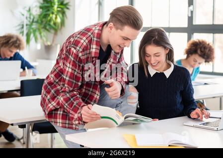 Friendly student helping his classmate to understand new information by explaining and showing her the answer in a textbook during break at college or Stock Photo