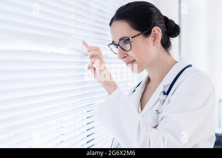 Portrait of a young female physician smiling while looking through the window during break in a modern and clean hospital Stock Photo