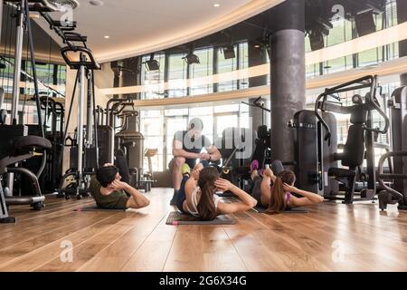 Fitness instructor timing three young people doing lateral crunch exercise with raised legs for abdominal muscles indoors at the gym Stock Photo