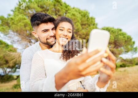 Young beautiful multiracial couple in love taking selfie in nature enjoying vacation moments sharing photos on social networks. New normal technology Stock Photo
