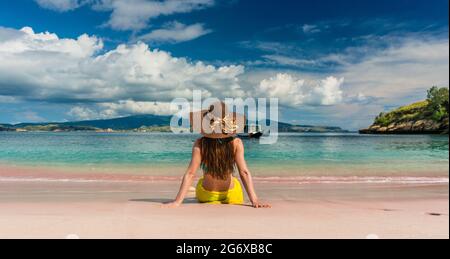 Rear view of a young woman with straw hat looking at an idyllic view while sitting on the sand at Pink Beach in Komodo Island, Indonesia Stock Photo