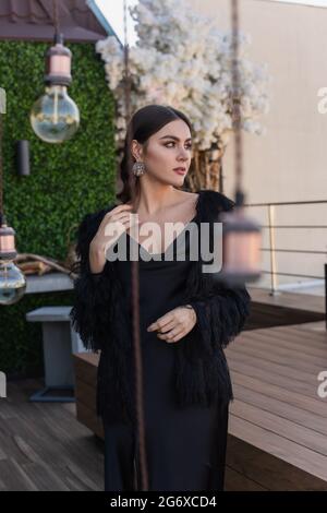 young woman in black slip dress and faux fur jacket looking away outside Stock Photo