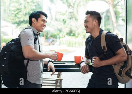 Side view of two cheerful young men wearing casual clothes and carrying trendy backpacks while talking and drinking coffee together in a cafeteria Stock Photo