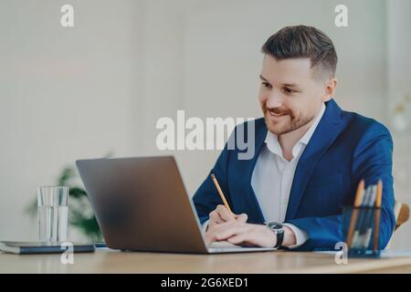 Corporate worker in formal suit sits at desktop focused at laptop computer poses in office interior writes down information with pencil watches Stock Photo