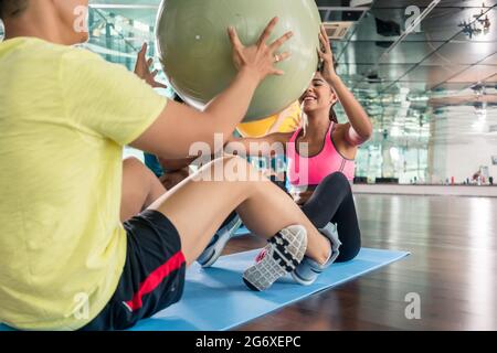 Cheerful young woman giving the fitness ball to her workout partner while doing crunches together for abdominal muscles in a modern health club Stock Photo