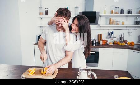 cheerful woman in white shirt covering eyes of man cutting orange in kitchen Stock Photo