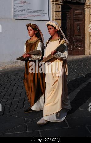 Two young ladies wearing typical Italian medieval costume carry decorative artworks as they walk through Piazza Guglielmo Marconi in Anagni, Lazio, Italy, in an historical August pageant recalling its past as City of the Popes, the summer retreat between the 11th and 14th centuries of several pontiffs.  The event also honours San Magno di Anagni, a 3rd century AD martyred bishop who became the town’s patron saint. Stock Photo