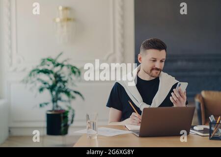 Male entrepreneur studying online, using smartphone while working at his workplace in modern office Stock Photo