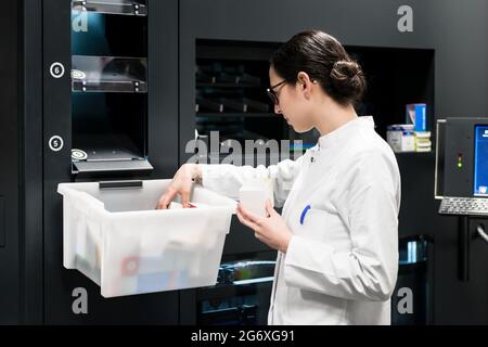 Side view of a dedicated female pharmacist choosing the best medicines from a plastic box during work in a contemporary drugstore with modern equipmen Stock Photo