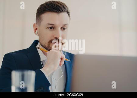 Professional male entrepreneur focused at laptop uses earphones listens necessary information watches webinar uses earphones holds pencil for writing Stock Photo