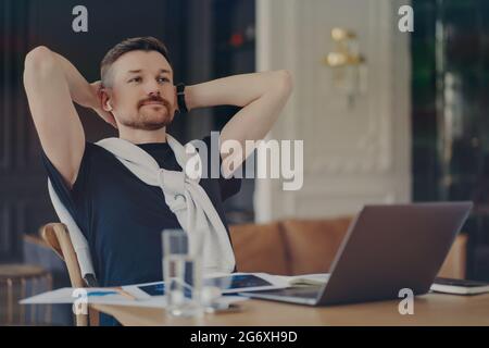 Pensive bearded man ponders on task keeps hands behind head sits at desktop with modern gadgets thinks about distance job concentrated into distance Stock Photo