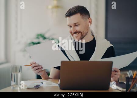 Handsome happy businessman analyzing documents and feeling satisfied while working on laptop from home Stock Photo
