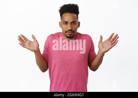 Confused african american guy shrugging clueless, raising hands sideways, empty arms, frowning complicated, cant understand, standing against white Stock Photo