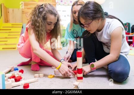 Two pre-school girls playing together with wooden toy blocks on the floor during playtime supervised by a careful young kindergarten teacher Stock Photo