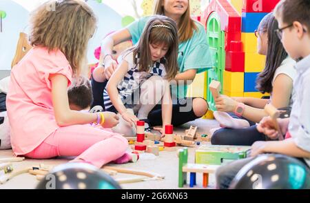 Dedicated young female kindergarten teacher holding a pre-school shy girl while watching children during playtime with wooden toy blocks in the classr Stock Photo