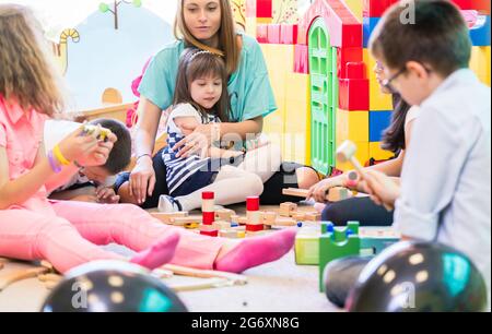 Dedicated young female kindergarten teacher holding a pre-school shy girl while watching children during playtime with wooden toy blocks in the classr Stock Photo