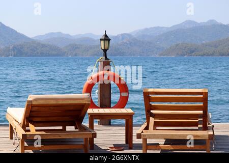Sea resort, two wooden deck chairs and lantern with lifebuoy on a pier on green mountains background. Beach vacation on picturesque nature Stock Photo