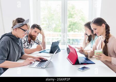 Dedicated young woman editing a document on laptop, while sharing the desk with other hard-working co-workers in a modern office space for digital nom Stock Photo