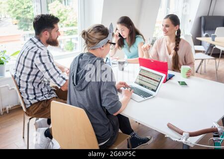 Dedicated young woman editing a document on laptop while sharing the desk, with other hard-working co-workers in a modern office space for digital nom Stock Photo