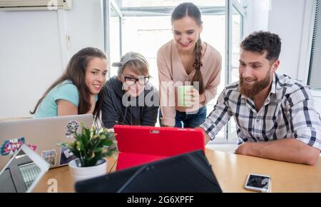 Four co-workers smiling, while watching together a professional business presentation or a funny video on tablet in a modern shared office space for f Stock Photo