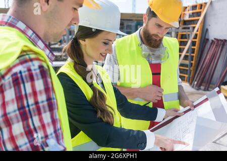 Young and confident female architect smiling while sharing ideas about the plan of a building under construction outdoors with two blue-collar workers Stock Photo
