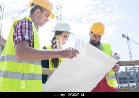 Experienced female architect smiling while analyzing a blueprint together with two workers outdoors on the construction site of a modern residential b Stock Photo