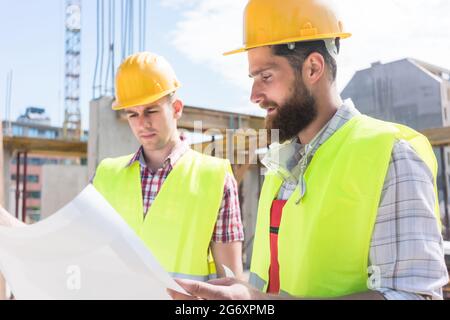 Two young construction workers wearing yellow hard hats and reflective safety vests while analyzing together the plan of a new building Stock Photo