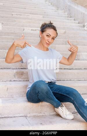 Middle aged women wearing t-shirt and jeans pointing on t-shirt sitting on the stone stairs outdoor Stock Photo