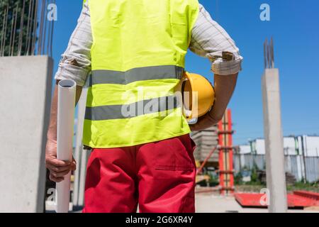 Midsection rear view of a worker holding a rolled blueprint and a yellow hard hat while standing on the construction site of a contemporary building Stock Photo