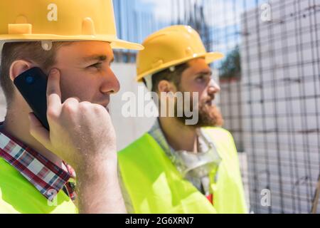 Side view close-up of a worker wearing yellow hard hat while talking on mobile phone during work, on the construction site of a building in progress Stock Photo