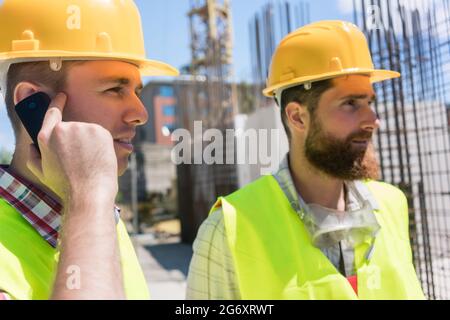 Side view close-up of a worker wearing yellow hard hat, while talking on mobile phone during work on the construction site of a building in progress Stock Photo