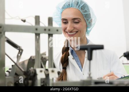 Portrait of a happy female technician writing a report while checking industrial equipment during quality control in a cosmetics factory Stock Photo