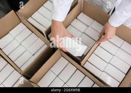 High-angle close-up view of the hands of a manufacturing worker putting packed products, in cardboard boxes before export or shipping during manual wo Stock Photo