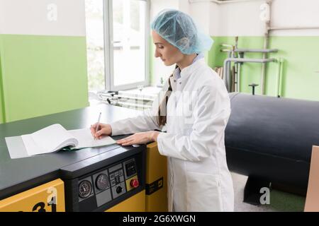 Side view of a female engineer writing observations for a report about equipment and production, while standing near a compressor machine in a modern Stock Photo