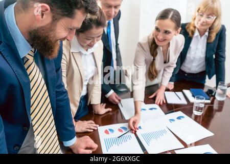 Multi-ethnic team of five dedicated specialists smiling while analyzing various charts with positive statistics about the profit and the development o Stock Photo