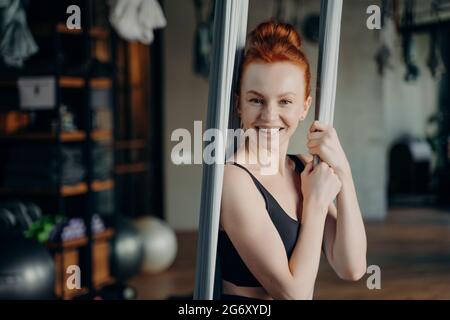 Excited red haired woman enjoying aerial yoga in fitness studio Stock Photo