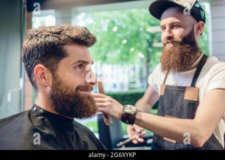 Dedicated male hairstylist using scissors and plastic comb while giving a cool haircut to a redhead bearded young man in a trendy beauty salon Stock Photo