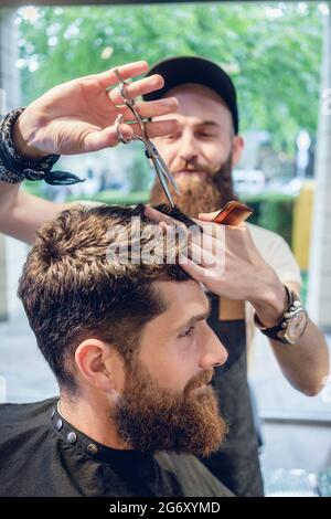 Dedicated male hairstylist using scissors and plastic comb while giving a cool haircut to a redhead bearded young man in a trendy beauty salon Stock Photo