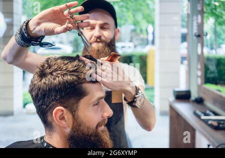 Dedicated male hairstylist using scissors and plastic comb while giving a cool haircut to a redhead bearded young man in a trendy beauty salon Stock Photo