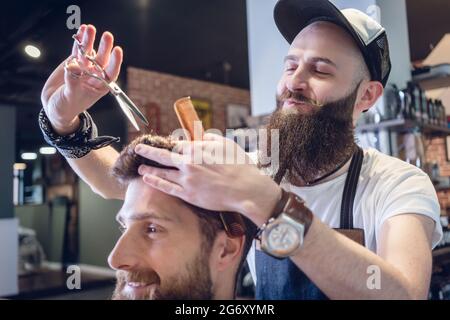 Dedicated male hairstylist using scissors and plastic comb while giving a cool haircut to a redhead bearded young man in a trendy beauty salon Stock Photo