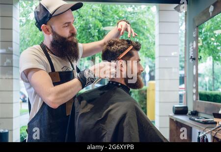 Dedicated male hairstylist using scissors and plastic comb while giving a cool haircut to a redhead bearded young man in a trendy beauty salon Stock Photo