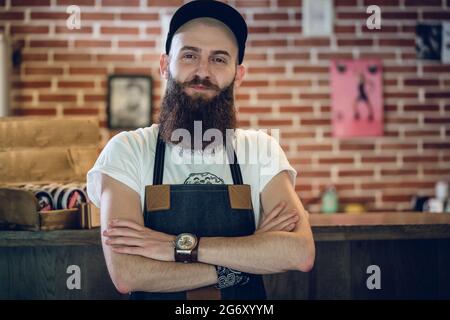 Portrait of a cool bearded hairstylist wearing wristwatch, apron and cap while looking at camera with confidence and passion for his job Stock Photo