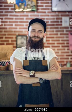 Portrait of a cool bearded hairstylist wearing wristwatch, apron and cap while looking at camera with confidence and passion for his job Stock Photo