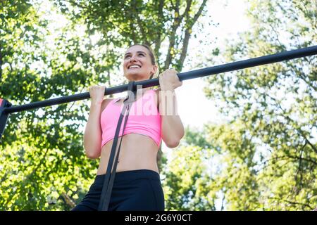 Low-angle view of a fit and cheerful young woman, wearing pink sports bra while doing chin-up exercise for the upper-body outdoors in a calisthenics p Stock Photo