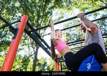Fit young woman smiling while doing pull-ups supported by her strong partner during couple workout routine in a calisthenics park in summer Stock Photo