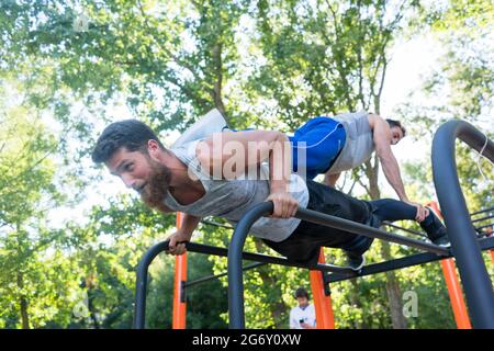 Low-angle view of a strong young man supporting his friend on his back, while doing extreme pull-ups during partner workout in a calisthenics park in Stock Photo