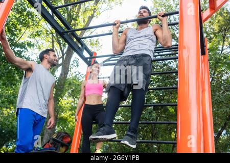 Low-angle view of a strong young man doing chin-up repetitions for the upper-body during calisthenics workout, with his friends in a modern outdoor fi Stock Photo